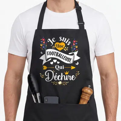 Tablier de cuisine noir avec inscription Je suis une footballeuse qui déchire entouré de fleurs et d'un cœur orange.