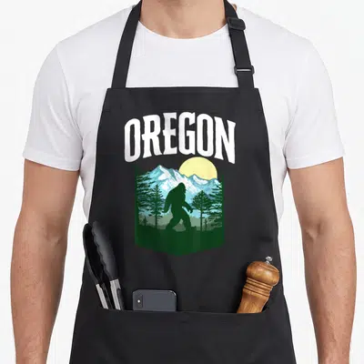 Tablier de cuisine noir avec silhouette Bigfoot, montagnes bleues et arbres verts sous le mot Oregon en blanc.