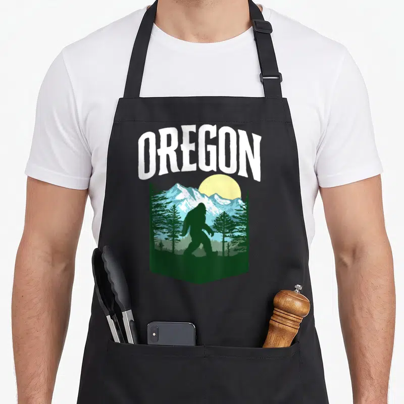 Tablier de cuisine noir avec silhouette Bigfoot, montagnes bleues et arbres verts sous le mot Oregon en blanc.