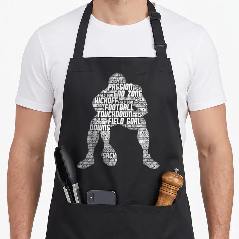 Tablier de cuisine noir avec silhouette de joueur de football américain composée de mots blancs comme sack et touchdown.