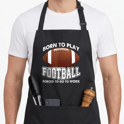 Tablier de cuisine noir avec une balle de football américaine et le texte Born to Play Football Forced to Go to Work en blanc.