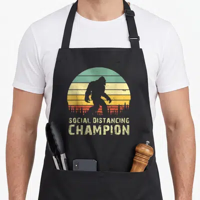 Tablier de cuisine noir avec silhouette de Bigfoot et texte Social Distancing Champion sur fond de soleil vintage.