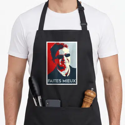 Tablier de cuisine noir avec un portrait graphique rouge et bleu portant l'inscription faites mieux en bas du motif imprimé.
