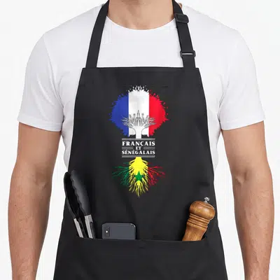 Tablier de cuisine noir avec drapeaux français et sénégalais fusionnés par un motif d'arbre blanc et racines colorées.