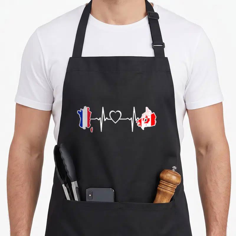 Tablier de cuisine noir avec carte de France et Canada reliées par un tracé de rythme cardiaque blanc et un cœur.