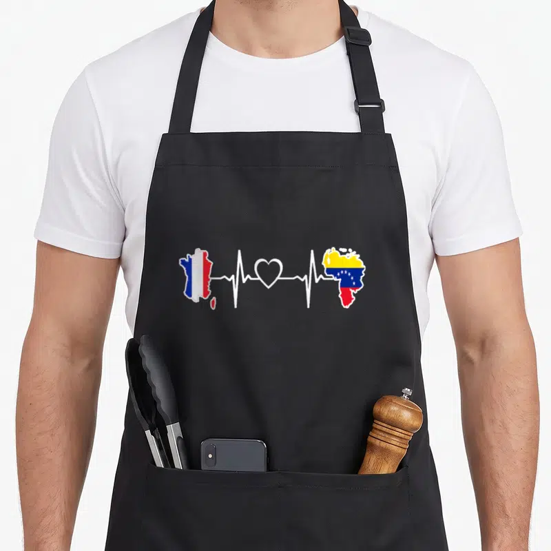 Tablier de cuisine noir avec carte France et Venezuela reliées par une ligne de rythme cardiaque blanche et un cœur.