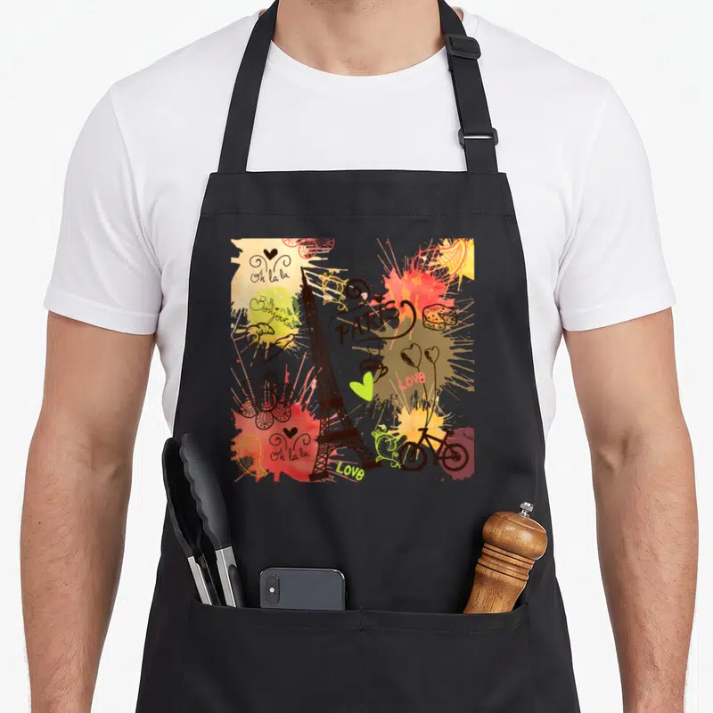 Tablier de cuisine noir avec une illustration colorée de Paris incluant la Tour Eiffel, un vélo et des textes comme Bonjour et Oh la la.