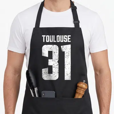 Tablier de cuisine noir avec l inscription blanche Toulouse 31 en typographie distressed et texturée.