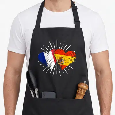Tablier de cuisine noir avec un cœur stylisé combinant les drapeaux français et espagnol entouré de traits blancs dynamiques.