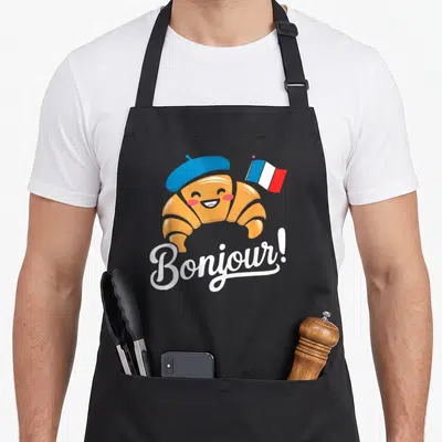 Tablier de cuisine noir avec un croissant souriant portant une béret bleue et un drapeau français, sous l'inscription Bonjour en blanc.