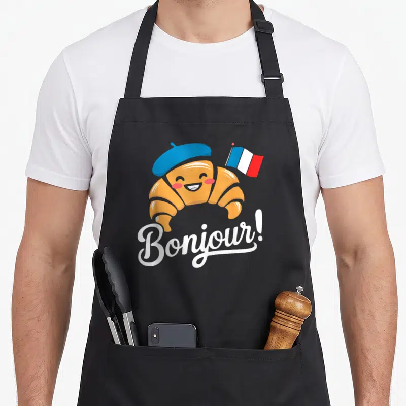 Tablier de cuisine noir avec un croissant souriant portant une béret bleue et un drapeau français, sous l'inscription Bonjour en blanc.