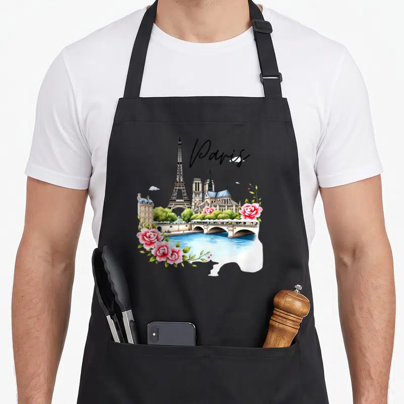 Tablier de cuisine noir avec illustration colorée de Paris incluant la Tour Eiffel, Notre-Dame, un pont sur la Seine et des roses roses.