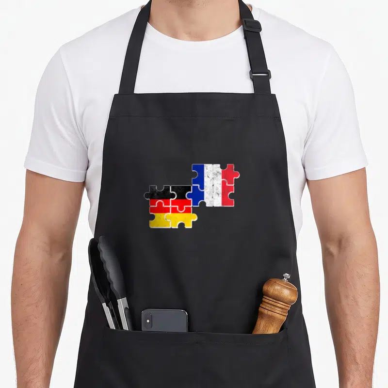 Tablier de cuisine noir avec drapeaux français et allemand en puzzle vintage superposés au centre.