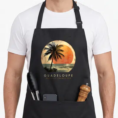 Tablier de cuisine noir avec illustration circulaire d'un palmier au coucher du soleil et texte Guadeloupe Antilles Françaises
