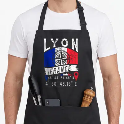 Tablier de cuisine noir avec drapeau français, boussole, skyline et coordonnées géographiques de Lyon imprimés en blanc et couleurs vives.