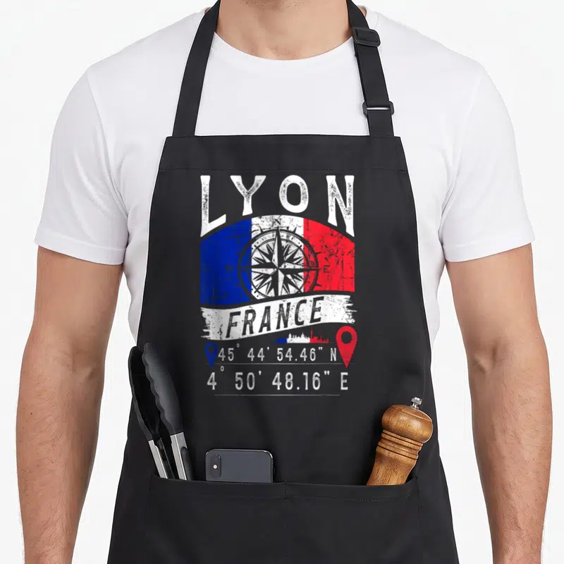 Tablier de cuisine noir avec drapeau français, boussole, skyline et coordonnées géographiques de Lyon imprimés en blanc et couleurs vives.