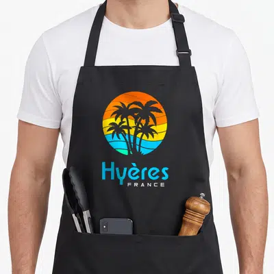 Tablier de cuisine noir avec un logo circulaire représentant des palmiers sur un fond de soleil orange et jaune au-dessus de l'eau bleue.