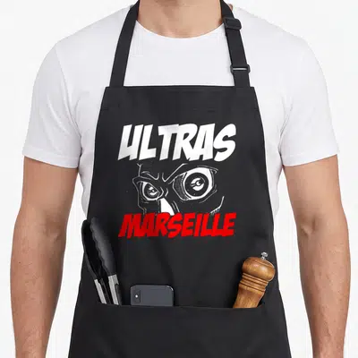 Tablier de cuisine noir avec inscription Ultras Marseille et illustration de visage aux yeux exagérés en blanc et rouge.