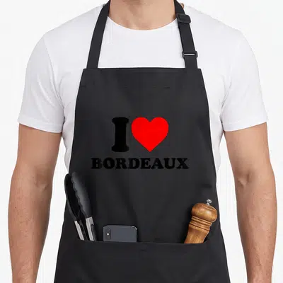 Tablier de cuisine noir avec un cœur rouge et une silhouette noire au-dessus du mot BORDEAUX en lettres capitales noires.