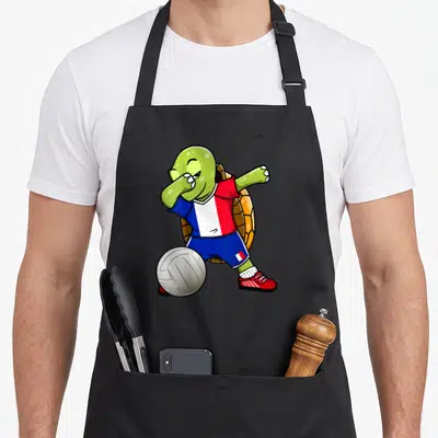 Tablier de cuisine noir avec une tortue verte en maillot français faisant un dab près d'un ballon blanc.