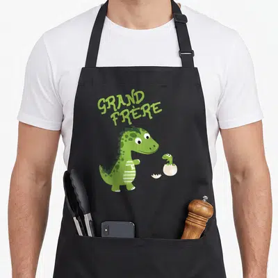 Tablier de cuisine noir avec un dinosaure vert souriant et un bébé dinosaure sortant d'une coque blanche sous le texte Grand Frère.