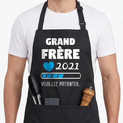 Tablier de cuisine noir avec texte Grand Frère 2021 Veuillez Patienter et icône cœur bleu sur fond uni.