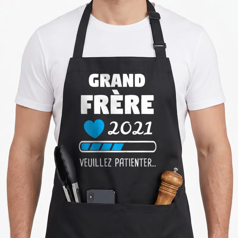 Tablier de cuisine noir avec texte Grand Frère 2021 Veuillez Patienter et icône cœur bleu sur fond uni.