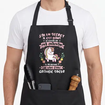 Tablier de cuisine noir avec licorne rose et texte annonçant la naissance d'un nouveau bébé dans la famille.
