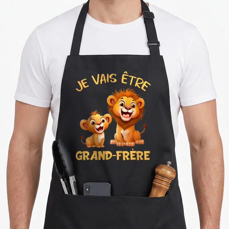 Tablier de cuisine noir avec deux lions colorés et le texte Je vais être grand-frère imprimé en jaune.