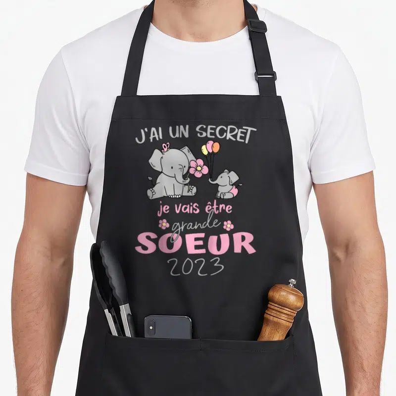 Tablier de cuisine noir avec deux éléphants roses, des fleurs et le texte annonce de naissance.