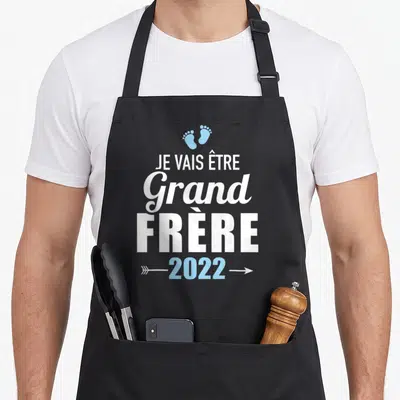 Tablier de cuisine noir avec inscription Je vais être grand frère 2022 et deux petits pieds bleus en haut du motif.