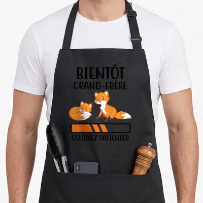 Tablier de cuisine noir avec deux renards orange et texte Bientôt grand-frère Veuillez patienter.