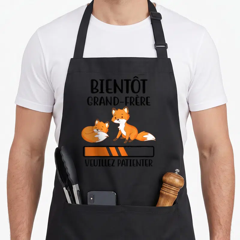 Tablier de cuisine noir avec deux renards orange et texte Bientôt grand-frère Veuillez patienter.