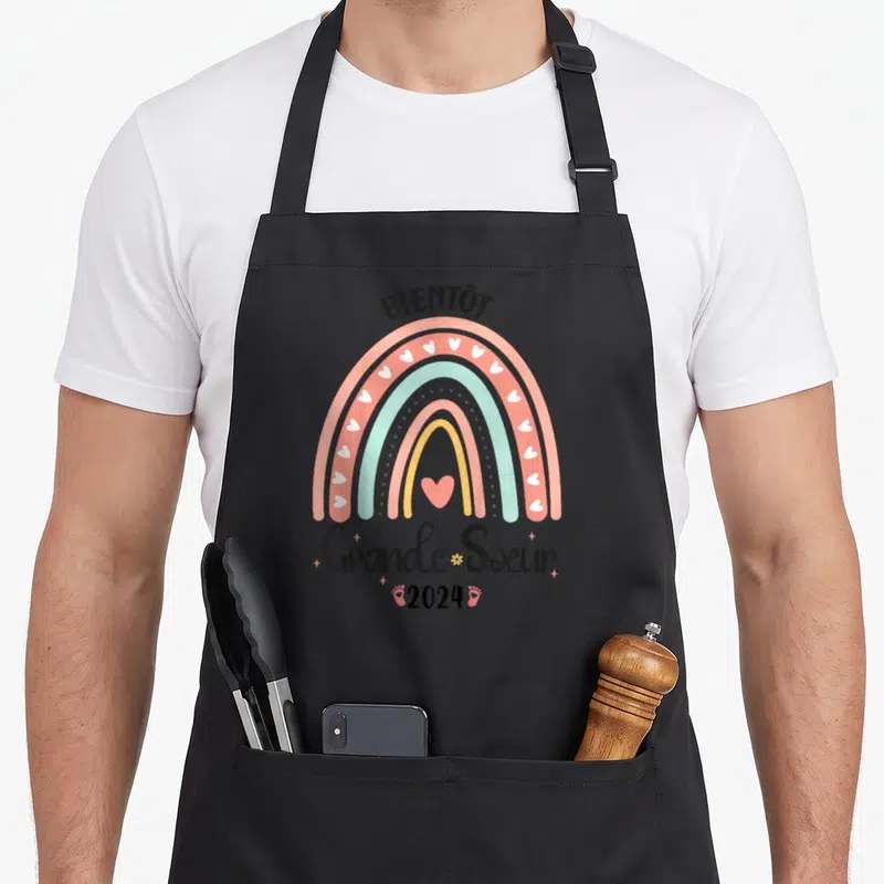 Tablier de cuisine noir avec arc-en-ciel pastel, cœurs blancs, inscription Bientôt Grande Soeur 2024 et petites empreintes de pieds roses en bas.