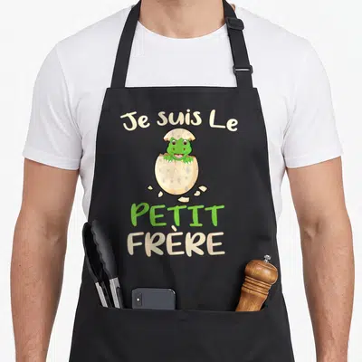Tablier de cuisine noir avec un bébé dinosaure vert sortant d'une coquille tachetée et du texte beige et vert.