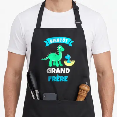Tablier de cuisine noir avec dinosaure vert et texte Bientôt Grand Frère imprimé en blanc et bleu.