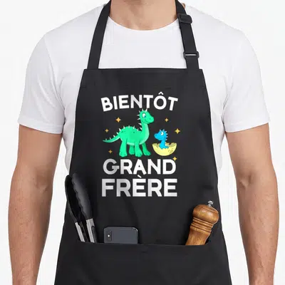 Tablier de cuisine noir avec un dinosaure vert et un bébé dinosaure sortant d'une coque jaune entourés d'étoiles.