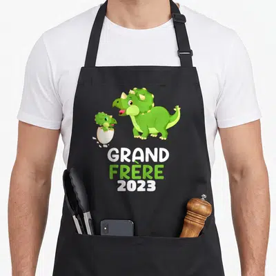 Tablier de cuisine noir avec deux dinosaures verts et l'inscription Grand Frère 2023 en lettres blanches et vertes.