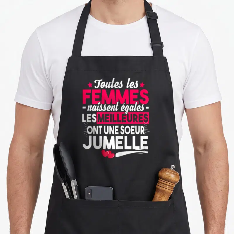 Tablier de cuisine noir avec texte rose et blanc célébrant les sœurs jumelles et deux petits cœurs rouges en bas.