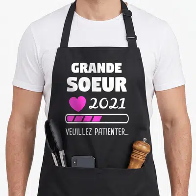 Tablier de cuisine noir avec inscription Grande Soeur 2021, cœur rose et barre de chargement.