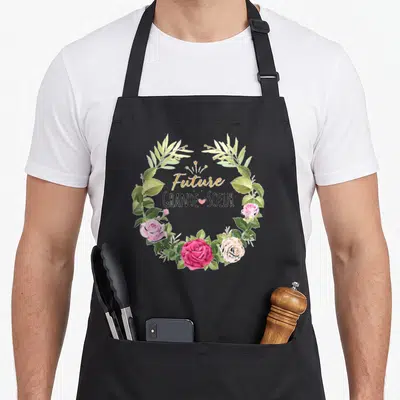 Tablier de cuisine noir avec une couronne de roses roses et de feuilles vertes autour du texte Future Grande Soeur.