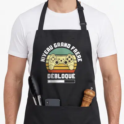 Tablier de cuisine noir avec illustration manette de jeu vidéo et texte Niveau Grand Frère Débloqué en style rétro.