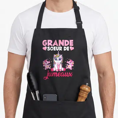 Tablier de cuisine noir avec une grande licorne rose entourée de deux petites licornes et des étoiles dorées.