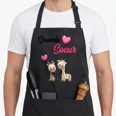 Tablier de cuisine noir avec deux girafes tenant des cœurs roses et l'inscription Grande Soeur en lettres noires et roses.