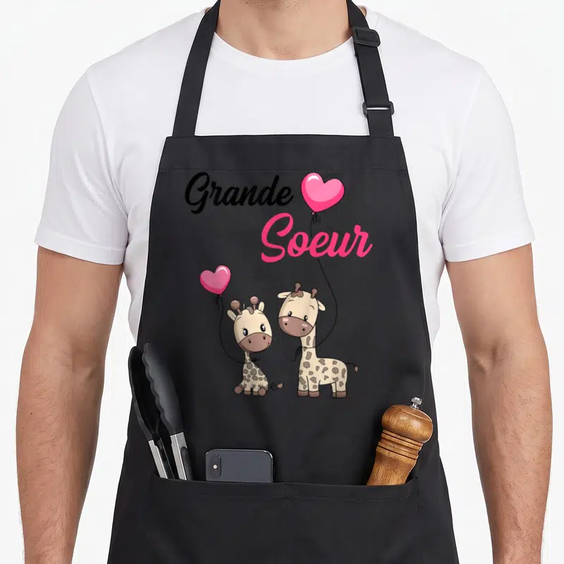Tablier de cuisine noir avec deux girafes tenant des cœurs roses et l'inscription Grande Soeur en lettres noires et roses.