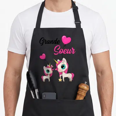 Tablier de cuisine noir avec deux licornes roses, des cœurs en forme de ballon et l'inscription Grande Soeur 2026.