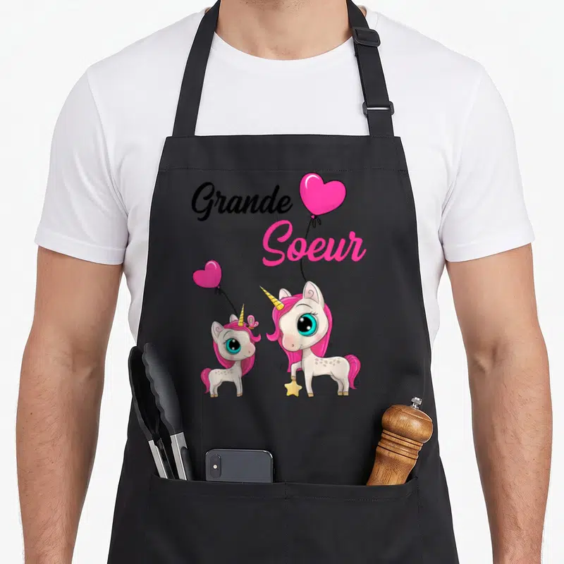 Tablier de cuisine noir avec deux licornes roses, des cœurs en forme de ballon et l'inscription Grande Soeur 2026.