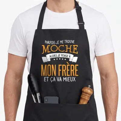 Tablier de cuisine noir avec inscription orange et blanche « Parfois je me trouve moche alors je pense à mon frère » sur fond gris uni.