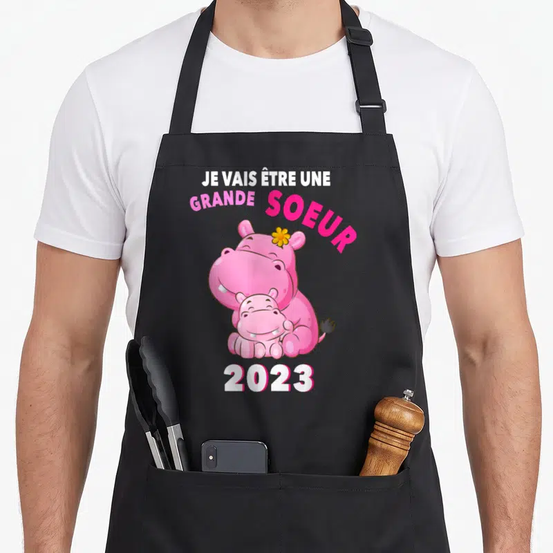 Tablier de cuisine noir avec deux hippopotames roses s'embrassant et le texte Je vais être une grande soeur 2023 imprimé en haut et en bas.