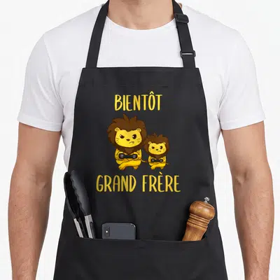Tablier de cuisine noir avec deux lions jaunes tenant des manettes et le texte Bientôt Grand Frère en jaune.